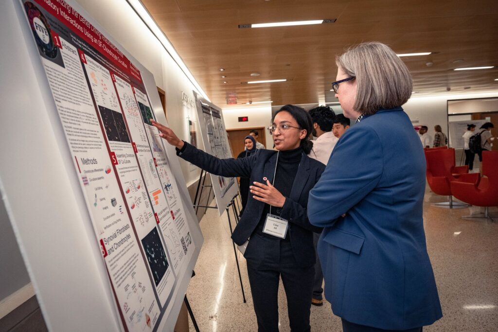 A student researcher explains her project board to a professor